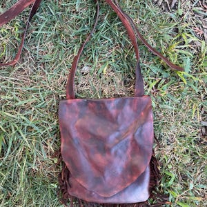 May include: A dark brown leather crossbody bag with a smooth, mottled finish and a pointed front flap. The bag features a long, adjustable leather shoulder strap and a decorative fringe trim along the bottom edge, creating a textured appearance.