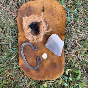 May include: A tan leather pouch containing a dark gray metal fire striker, a light gray flint stone, a small circular white disc, and a bundle of fibrous brown tinder. The items are arranged on the surface of the leather pouch.