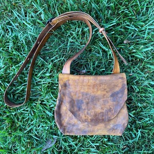 May include: A brown leather crossbody bag with a flap closure and an adjustable shoulder strap. The bag features a distressed, mottled texture with variations in color ranging from light tan to dark brown across the surface of the leather.
