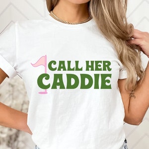 May include: White t-shirt with a pink golf flag and green text that reads "Call Her Caddie".