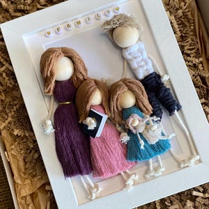 May include: A white frame with the word "FAMILY" spelled out in gold letters. Inside the frame are five yarn dolls, two with long brown hair, one with short brown hair, one with long blonde hair, and one with short blonde hair. The dolls are all wearing different colored clothing.