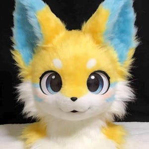 May include: A plush fox head with large, expressive eyes. The fox features yellow fur with blue accents on the ears and cheeks. The eyes are large and round with blue irises. The fox has a white muzzle and a small, black nose.
