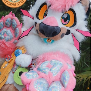 May include: A white and pink furry fursuit head and paws. The head has large orange and blue eyes, a pink nose, and a pink mohawk. The paws are pink with iridescent paw pads. The logo "Fursuit Design PPKBunny Studio" is visible.