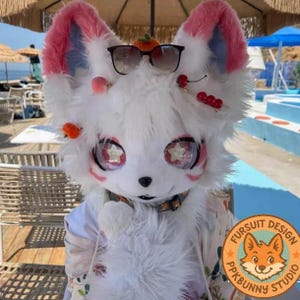 Può includere: Una testa di fursuit bianca con orecchie foderate di rosa, grandi occhi con disegni a stella e un naso nero. Il fursuit ha occhiali da sole sopra ed è decorato con piccoli accessori di frutta. Un logo con una volpe è visibile in basso a destra.