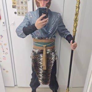 Puede incluir: Una persona vestida como el Rey Mono de Viaje al Oeste. Lleva una t&uacute;nica gris y azul con una falda de piel de tigre. Sostiene un bast&oacute;n largo y negro con una punta dorada.