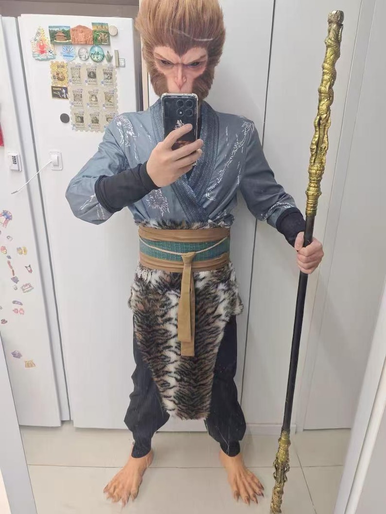 Puede incluir: Una persona vestida como el Rey Mono de Viaje al Oeste. Lleva una t&uacute;nica gris y azul con una falda de piel de tigre. Sostiene un bast&oacute;n largo y negro con una punta dorada.