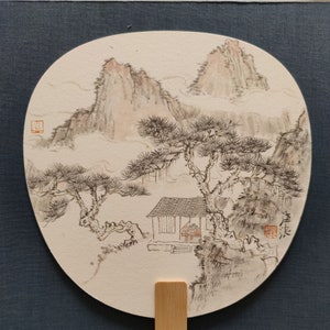 Puede incluir: Un abanico chino tradicional con una escena pintada de un paisaje de montaña con una pequeña casa y árboles. El abanico está hecho de papel y tiene un mango de bambú.