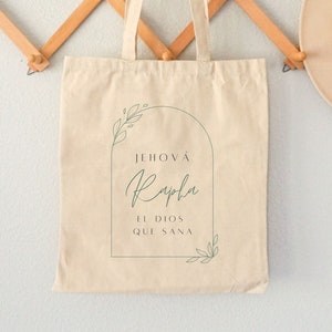 May include: A beige canvas tote bag with a green floral design and the text "Jehová Rapha El Dios Que Sana" printed on the front.