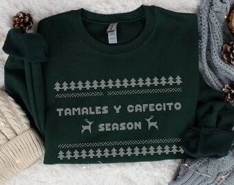 May include: A dark green sweatshirt with white embroidered Christmas tree and reindeer designs. The text "TAMALES Y CAFECITO SEASON" is displayed in white. The sweatshirt is a cozy, festive garment.