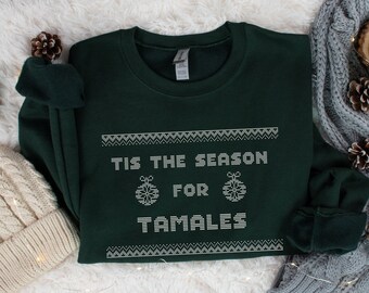 May include: A dark green sweatshirt with the text "TIS THE SEASON FOR TAMALES" in white, along with decorative snowflake ornaments. The sweatshirt is displayed on a white, fluffy surface, with a gray knitted scarf and pine cones in the background.
