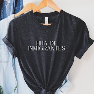 May include: A dark grey t-shirt with the text "HIJA DE INMIGRANTES" printed in white on the front.