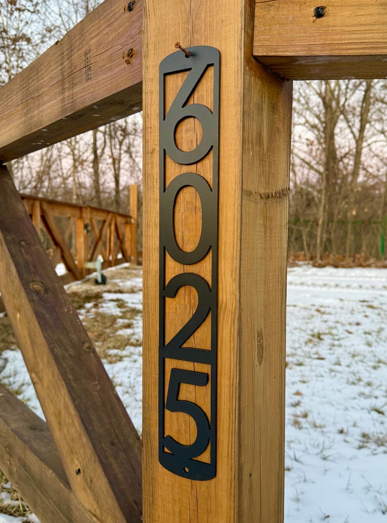 May include: Black metal house number plaque with the numbers 6025 attached to a wooden fence post.