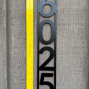 May include: Black metal house number plaque with the numbers 2025 cut out. The plaque is 20 inches tall and is shown next to a ruler.