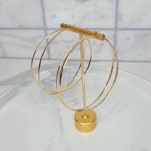 May include: A pair of large, thin, gold-toned hoop earrings suspended on a gold-colored metal T-bar display stand. The earrings feature a simple, circular wire design with a polished metallic finish, hanging vertically from the horizontal bar.