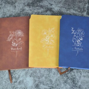 Op de afbeelding: Drie lederen notitieboekjes in bruin, geel en blauw met bloemmotieven en de namen "Rachel", "Chelsea" en "Natalie" gegraveerd op de voorzijden.