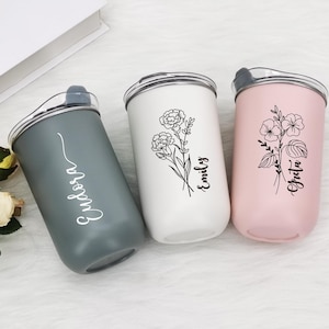 Könnte beinhalten: Drei personalisierte Becher in Grau, Weiß und Rosa. Jeder Becher hat ein Blumendesign und einen Namen in Schreibschrift. Die Becher haben einen silbernen Rand und einen transparenten Deckel. Der graue Becher hat den Namen "Eudora". Der weiße Becher hat den Namen "Emily". Der rosa Becher hat den Namen "Greta".