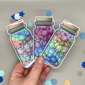 Pode incluir: Três autocolantes de frascos cheios de estrelas de origami coloridas. Os frascos têm um efeito holográfico, com um brilho de arco-íris. As estrelas no interior são tons de rosa, azul e amarelo. Os frascos têm uma tampa de cortiça e são rodeados por confetes em forma de estrela.