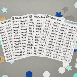 Pode incluir: Uma coleção de folhas de adesivos brancos com texto preto, apresentando os números das semanas de 1 a 52. Os adesivos são rotulados como "Tablet & Quill". O fundo é cinza com confetes azuis e brancos.