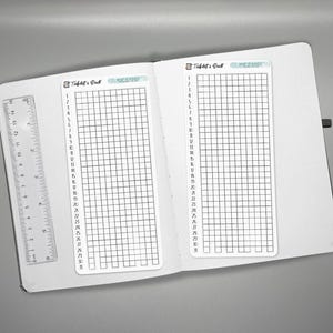 May include: Open white notebook with two habit tracker inserts and a clear ruler. Each tracker has a grid of boxes with numbers 1-30 down the side. The top of each tracker reads "Habit & Goal" and "MEANS".