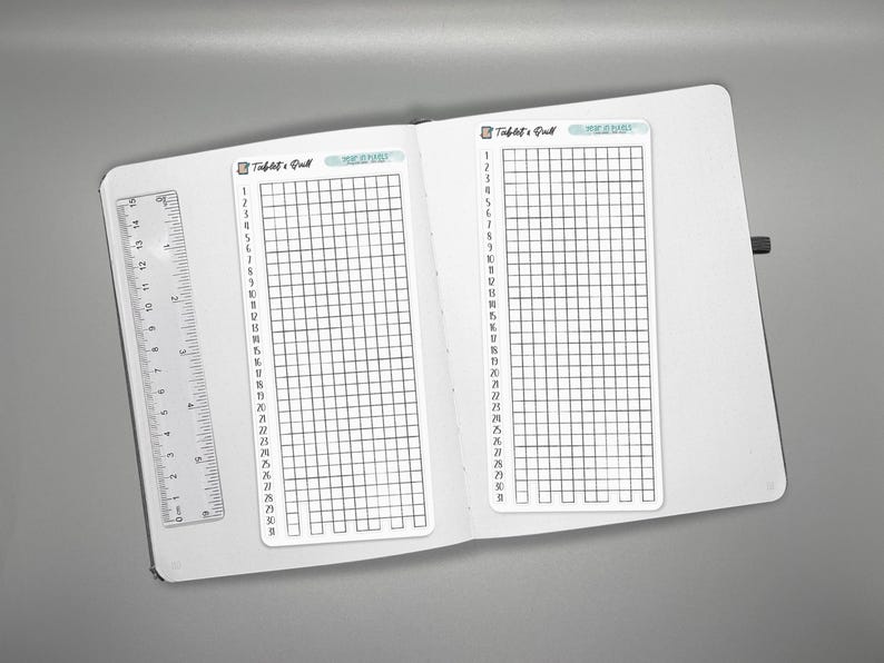 May include: Open white notebook with two habit tracker inserts and a clear ruler. Each tracker has a grid of boxes with numbers 1-30 down the side. The top of each tracker reads "Habit & Goal" and "MEANS".