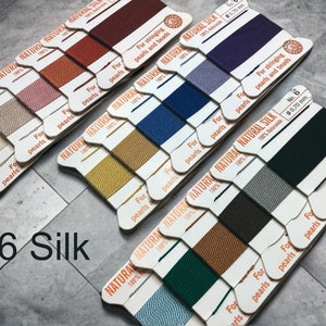 May include: A collection of spools of natural silk thread in various colors, including red, purple, and green. Each spool is labeled with "NATURAL SILK" and "For stringing pearls and beads." The text "#6 Silk" is also visible.