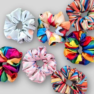 May include: A selection of scrunchies in a variety of vibrant patterns. The scrunchies feature designs such as rainbow stripes, floral prints, and abstract art. These hair accessories are made from a silky material and are in a range of colours.