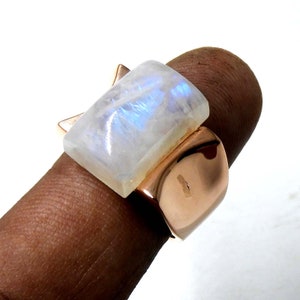 May include: A ring with a large, rectangular, white moonstone set in a rose gold band. The ring is worn on a finger.