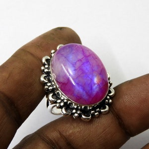 May include: A silver ring featuring a large, oval-shaped gemstone with a vibrant purple and pink color gradient. The gemstone is set in a decorative, ornate silver setting with a floral design. The ring is shown on a person's finger.