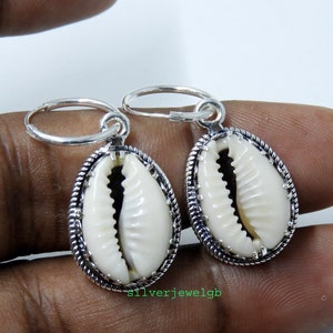 Peut inclure: Une paire de boucles d'oreilles créoles en argent avec un pendentif en coquille de cauris blanc. La coquille est entourée d'une bordure argentée avec un motif en forme de corde.