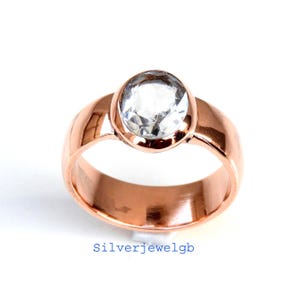 May include: A rose gold ring featuring a large, clear, oval-cut gemstone. The ring has a wide band and a polished finish. The text "Silverjewelgb" is printed in blue at the bottom of the image.