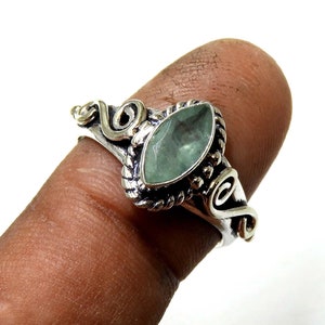 Puede incluir: Un anillo de plata con una piedra preciosa verde pálido en forma de marquesa. El anillo presenta detalles ornamentados alrededor de la piedra y en la banda, con un diseño en espiral. El anillo se muestra en un dedo.