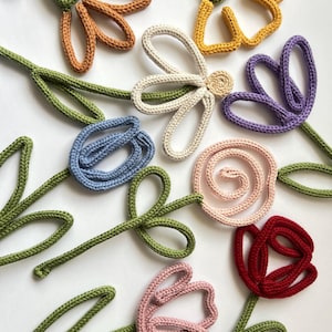 Puede incluir: Una colección de flores de ganchillo en varios colores, incluyendo verde, azul, morado, rosa, rojo, naranja y amarillo. Las flores son todas de diferentes formas y tamaños, y están dispuestas en un patrón aleatorio.