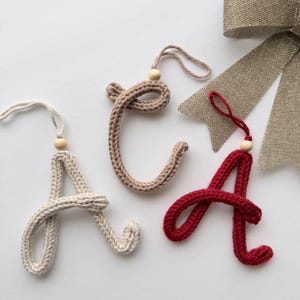May include: Three handmade crochet letter "A" ornaments in cream, tan, and red, each with a wooden bead and string for hanging. A gold ribbon bow is in the background. These decorative letters are perfect for holiday decor.