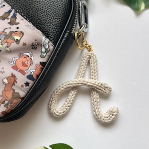May include: A white crocheted letter A keychain attached to a black leather purse with a cartoon print. The purse has a gold chain and a gold clasp.