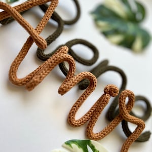May include: The word "hello" spelled out in brown and green yarn. The yarn is woven in a knit stitch. The letters are on a white background.