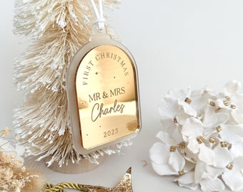 May include: A gold-colored Christmas ornament with the text "FIRST CHRISTMAS" and "MR & MRS Charles 2023". The ornament is hanging from a white string. A small white Christmas tree and white flowers are in the background.