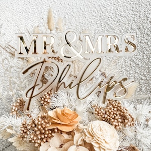 May include: A white acrylic cake topper with gold lettering that reads "MR & MRS Phillips". The topper is surrounded by dried flowers in shades of white, beige, and brown.