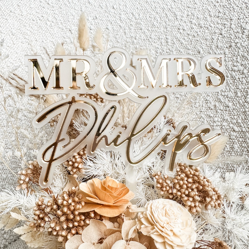May include: A white acrylic cake topper with gold lettering that reads "MR & MRS Phillips". The topper is surrounded by dried flowers in shades of white, beige, and brown.
