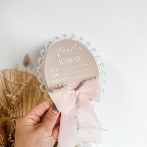 Peut inclure: Un panneau de naissance ovale beige avec un bord festonné et un ruban rose. Le panneau porte le nom "Amalia KARAS" et des espaces pour la date, l'heure, le poids et la taille. Le panneau est tenu par une main.