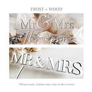 May include: A frosted acrylic cake topper with the text "Mr & Mrs Thompson" in a cursive font. The topper is on a white background with dried flowers.