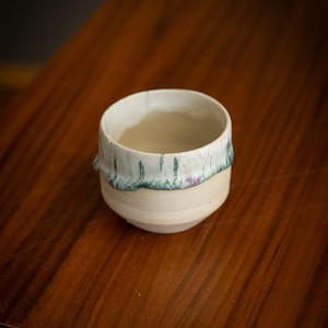 Op de afbeelding: Een kleine, handgemaakte keramische kom met een crèmekleurige basis en een wit interieur. De rand heeft een decoratieve band van groene en paarse glazuur. De kom staat op een houten oppervlak.