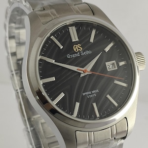 May include: A stainless steel Grand Seiko wristwatch with a black dial and silver hands. The watch features a date display and a silver link bracelet. The dial has the text "Grand Seiko" and "Spring Drive 5 Days".