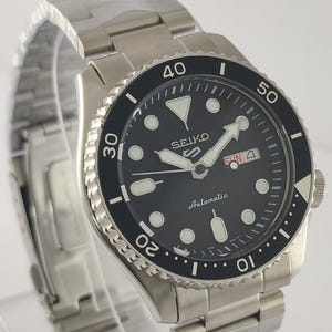 May include: A stainless steel Seiko 5 automatic watch with a black dial and a rotating bezel. The watch face displays the day and date, with luminous hands and markers. The watch has a stainless steel strap.