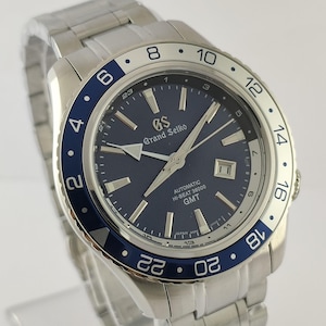 May include: A Grand Seiko wristwatch with a dark blue dial, silver-tone hands, and indices. The watch features a stainless steel bracelet, a date window at the three o'clock position, and a rotating bezel with a blue and silver 24-hour GMT scale.