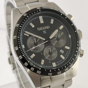 Op de afbeelding: Een Seiko chronograaf horloge van roestvrij staal met een zwarte wijzerplaat. Het horloge heeft meerdere sub-wijzerplaten, een tachymeter bezel en een roestvrijstalen armband. De wijzerplaat toont het Seiko-logo en diverse markeringen.