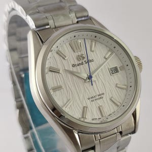 May include: A silver-toned stainless steel wristwatch with a textured white dial, featuring the Grand Seiko logo, a blue second hand, a date window at the three o'clock position, and text reading HI-BEAT 36000 80 HOURS and JAPAN.