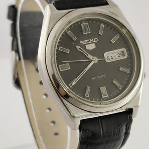 Może przedstawiać: Zegarek automatyczny Seiko 5 w stylu vintage z czarną tarczą i czarnym skórzanym paskiem. Zegarek ma srebrną kopertę i wyświetla dzień i datę. Tarcza zegarka ma prostokątne znaczniki godzin i tekst "SEIKO" i "AUTOMATIC".