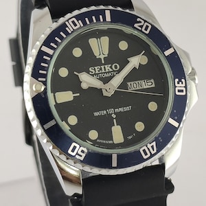 May include: A black Seiko automatic wristwatch with a blue rotating bezel. The watch face is black with white markers and hands, and the day is displayed as "MON 15". The watch has a black rubber strap.