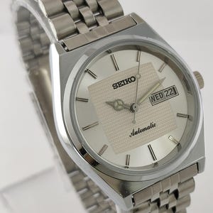 May include: Silver-toned Seiko automatic wristwatch with a rectangular face. The watch features a silver dial with a textured square center, black hour markers, and a day-date display. The band is a matching silver link bracelet.