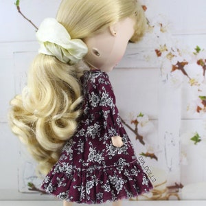 May include: A doll with long blonde hair styled in a ponytail with a white scrunchie. The doll is wearing a burgundy dress with a white floral pattern and a ruffled hem. The doll is standing on a white surface.